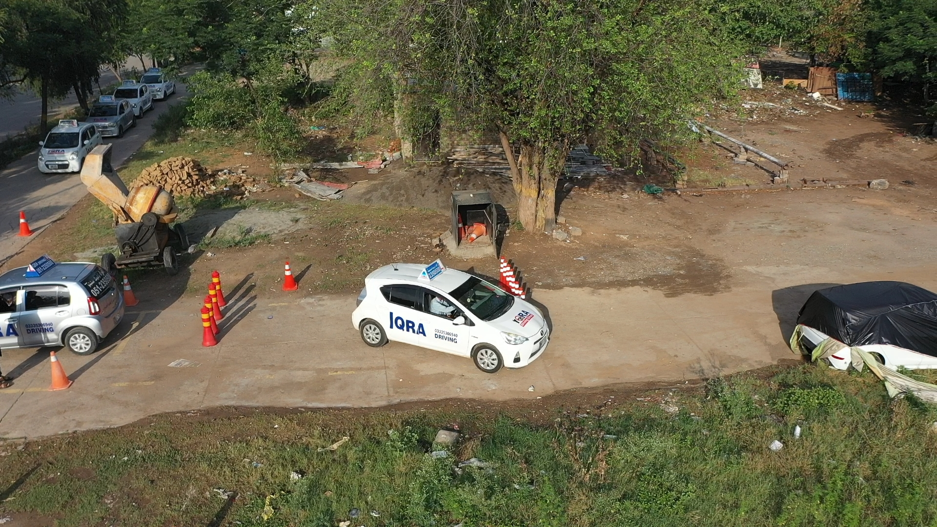 Aerial view of a driving school training area with white learner cars, orange traffic cones, and a practice course surrounded by trees.